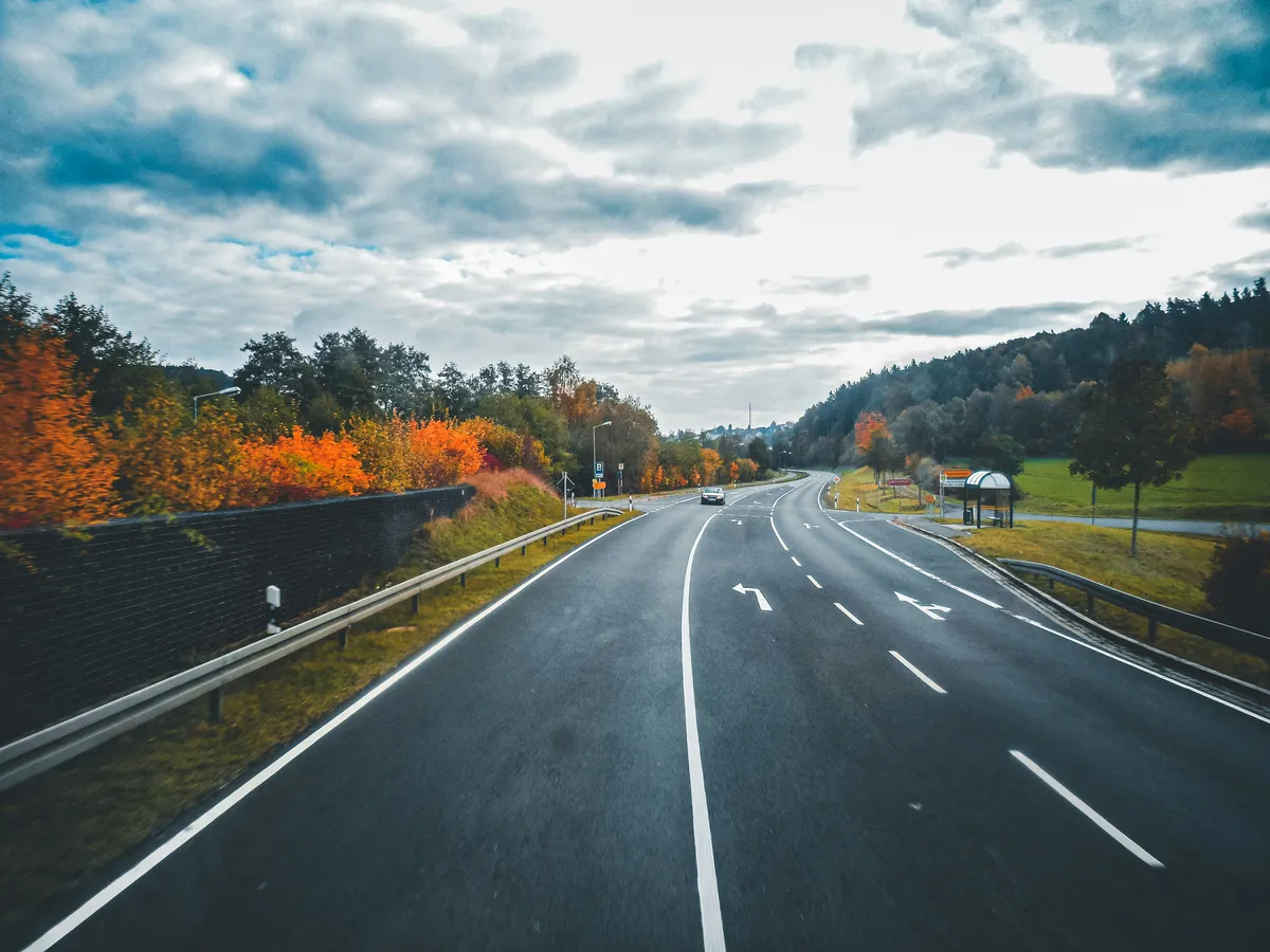 A motorway bordered by orange autumn trees beneath a cloudy sky