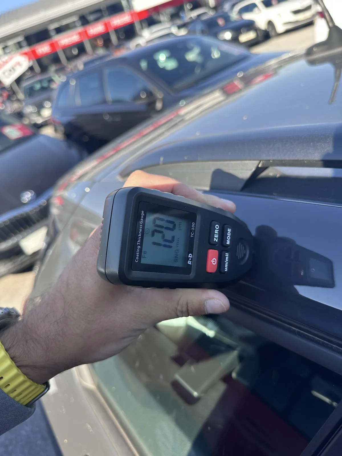 A paint thickness gauge showing 120 micrometres on a car panel during an outdoor inspection