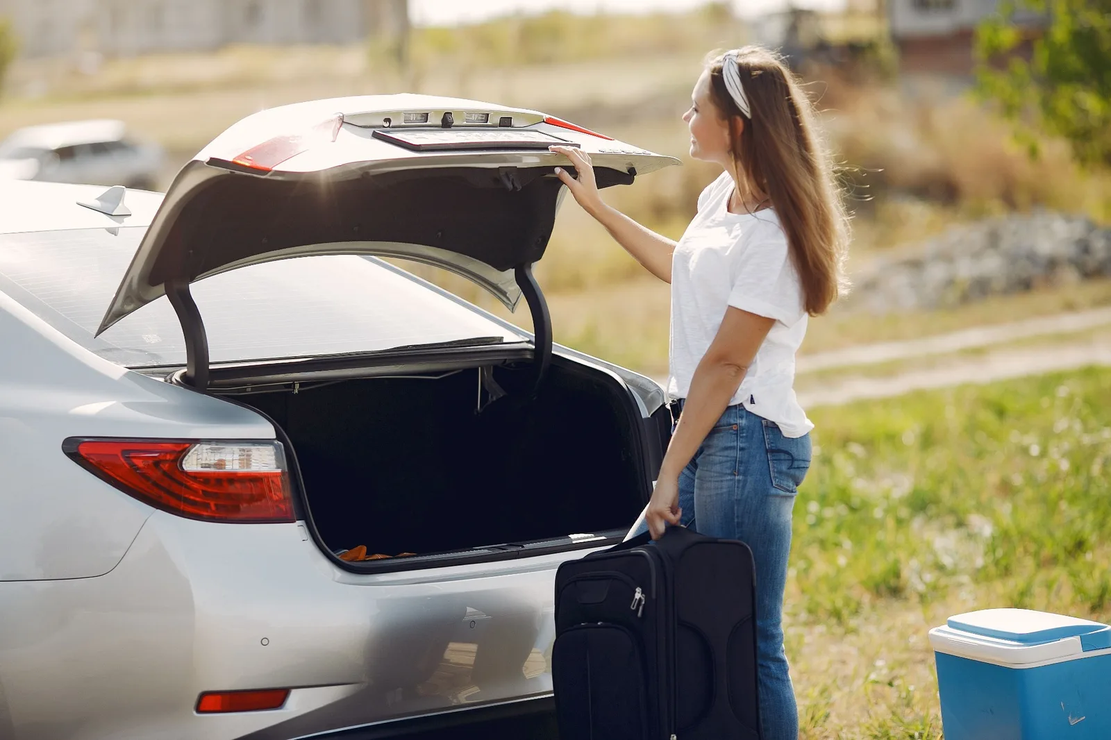 A woman with a suitcase opening the boot of a silver car before a trip
